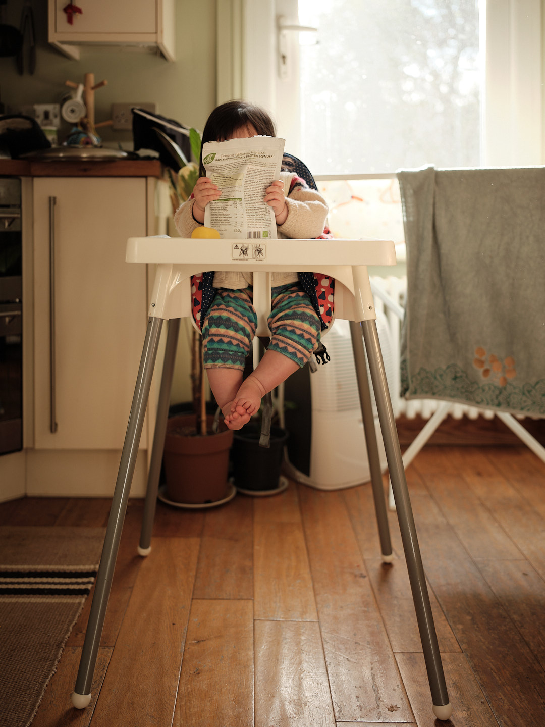 A baby sitting in a high chair in a kitchen, holding a bag of some cooking ingredients in front of her face.