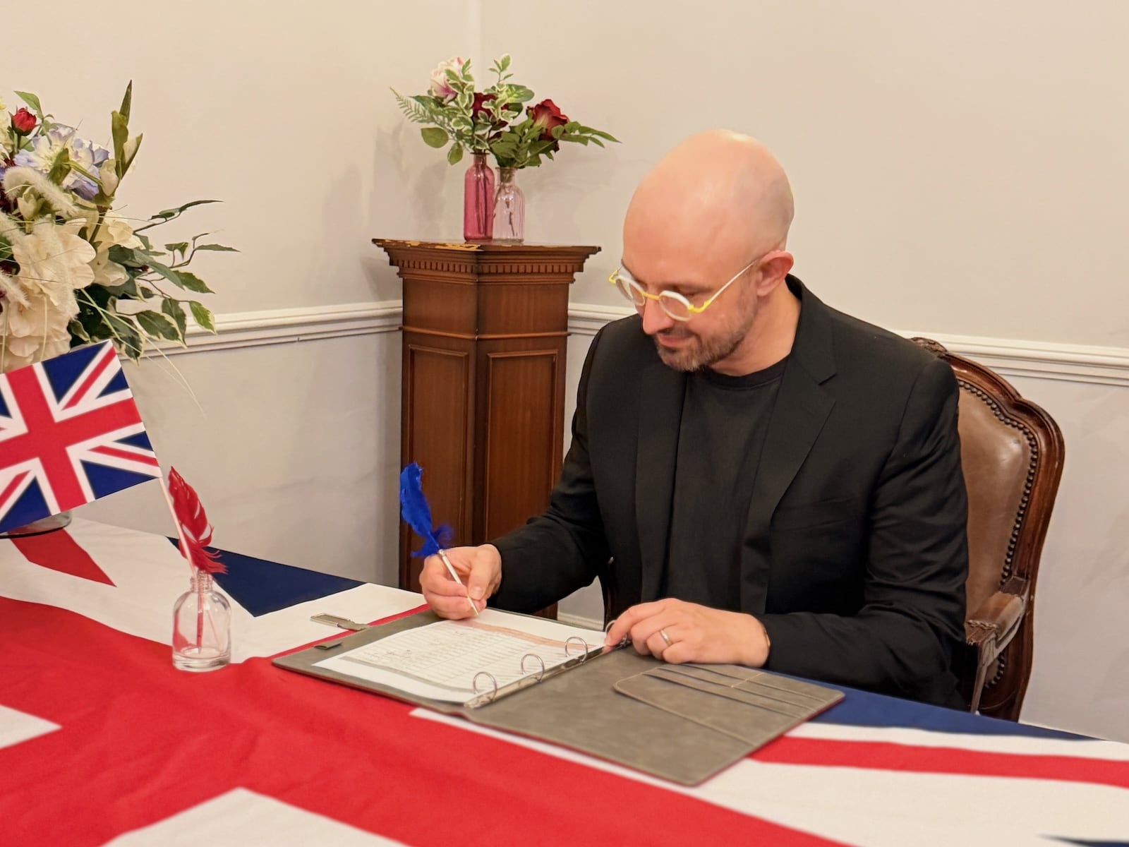 The author sitting at a desk covered with a British flag, holding a quill and apparently signing something