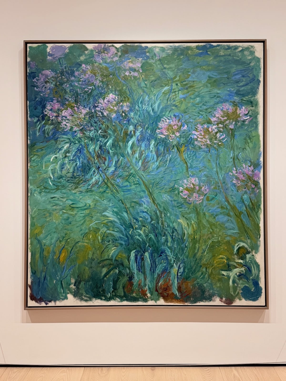 A photograph of a large square painting by Monet of agapanthus flowers: clusters of pale purple blooms on tall stems over a swirling mass of green and blue brushstrokes