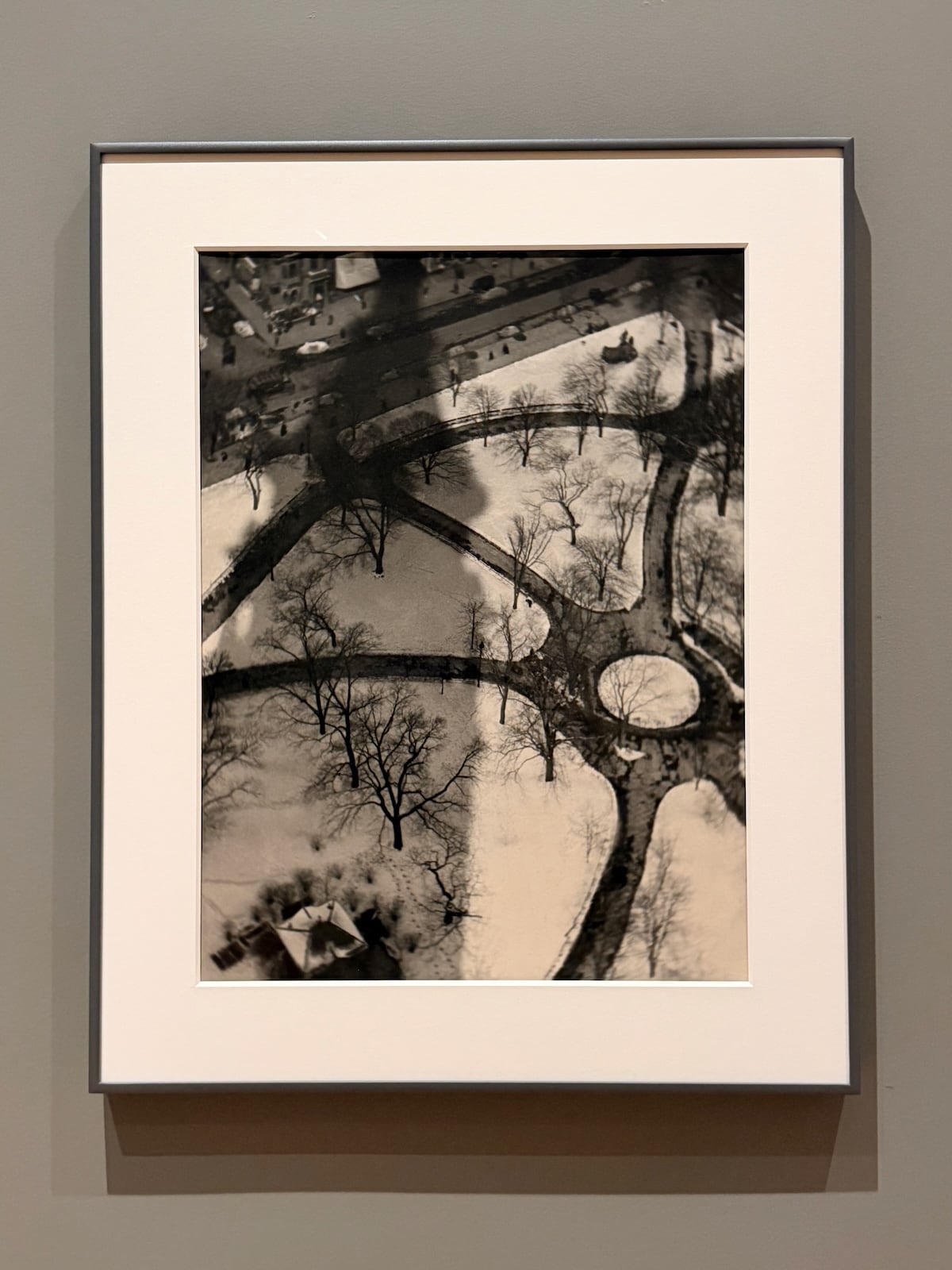 A photograph of a framed black and white photograph taken from high above a snow-covered park, showing dark, curving paths and bare trees radiating out from a central circle, with the long shadow of a tall building falling across the snow