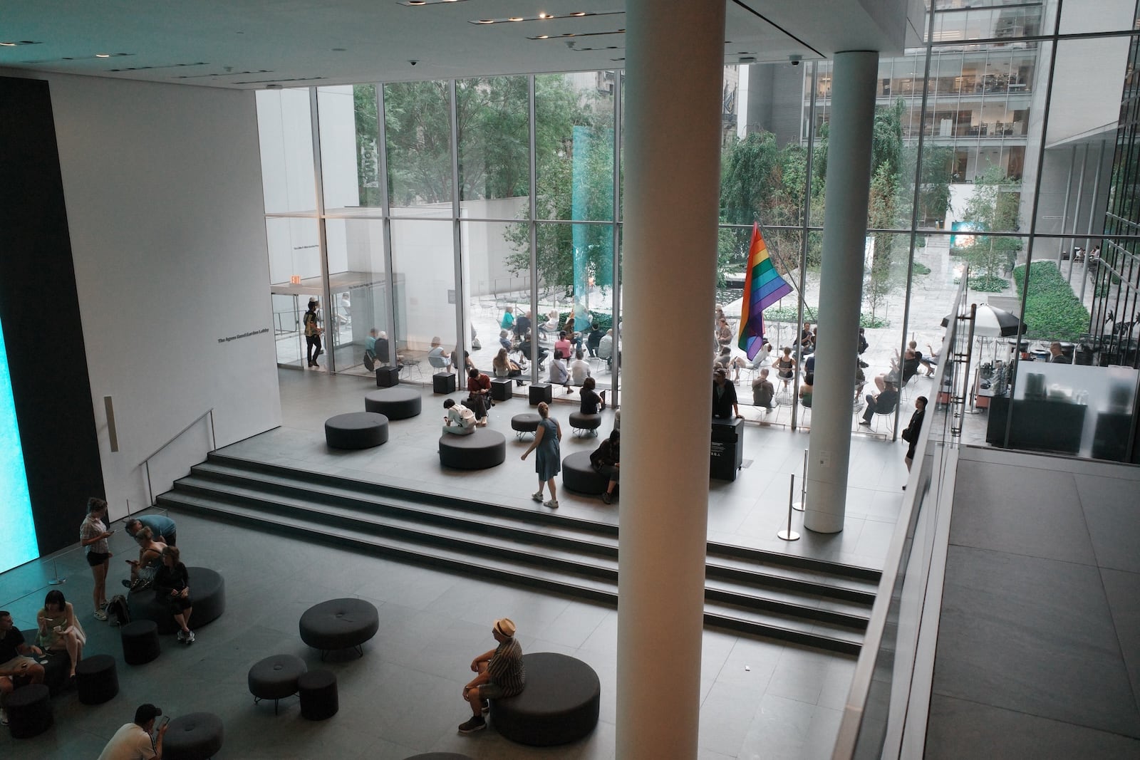 A photograph of an open space inside the MoMA, with people lounging on round seats. A tall, wide window separates the space from a courtyard, which includes more seating and some trees