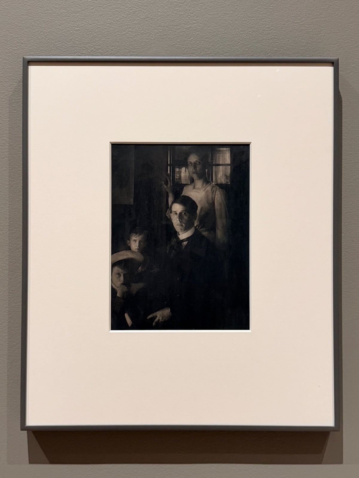 A photograph of a framed, dark-toned early 20th century photographic print showing a family: a man in a dark suit seated in the centre, a woman standing behind him by a window, and two children in straw hats in the foreground