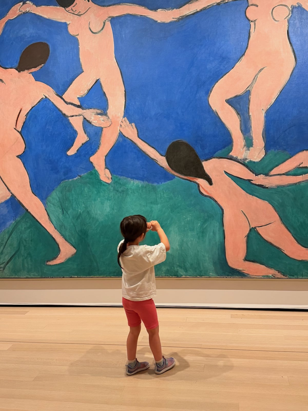 A photograph of a small child with a ponytail, in a white t-shirt and pink shorts, standing with a hand raised to her face in front of Matisse's huge painting 'Dance (I)', showing five pink figures dancing in a circle on a green hill against a blue sky