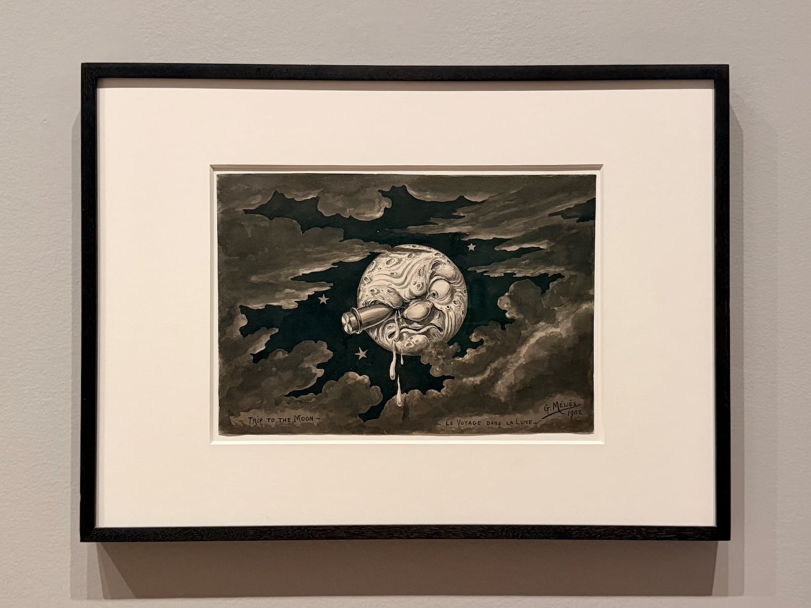 A photograph of a framed ink drawing of the moon with a grimacing human face and a rocket lodged in its eye, surrounded by dark clouds and stars, captioned ‘Trip to the Moon’ and ‘Le Voyage dans la Lune’ and signed G. Méliès 1902