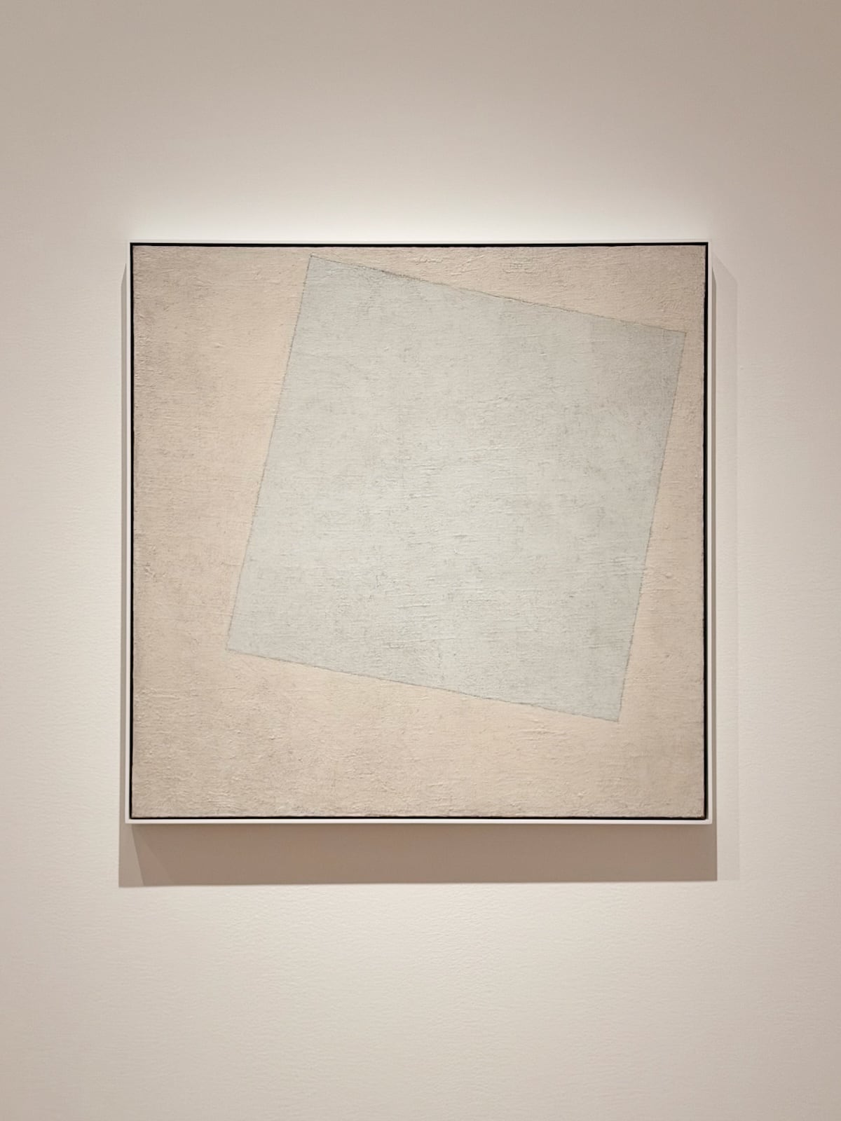 A photograph of a square painting on a beige wall: a tilted off-white square on a slightly warmer white background, with visible brushstrokes and cracking in the paint
