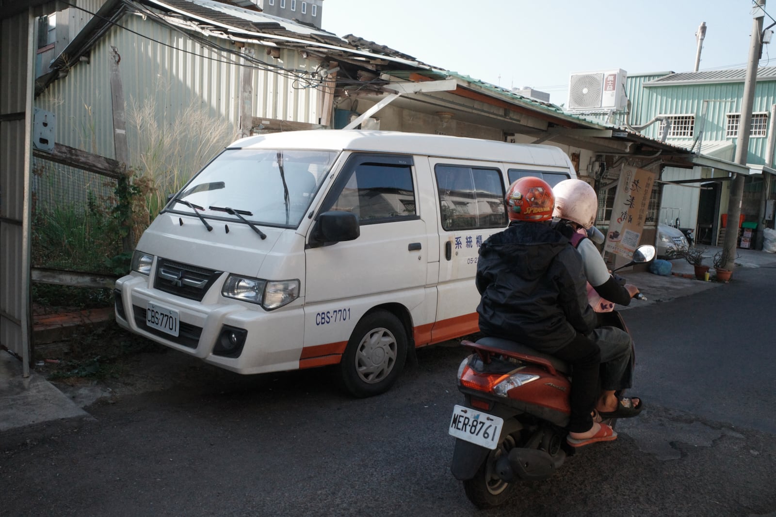 Two people riding a scooter past a parked Delica van
