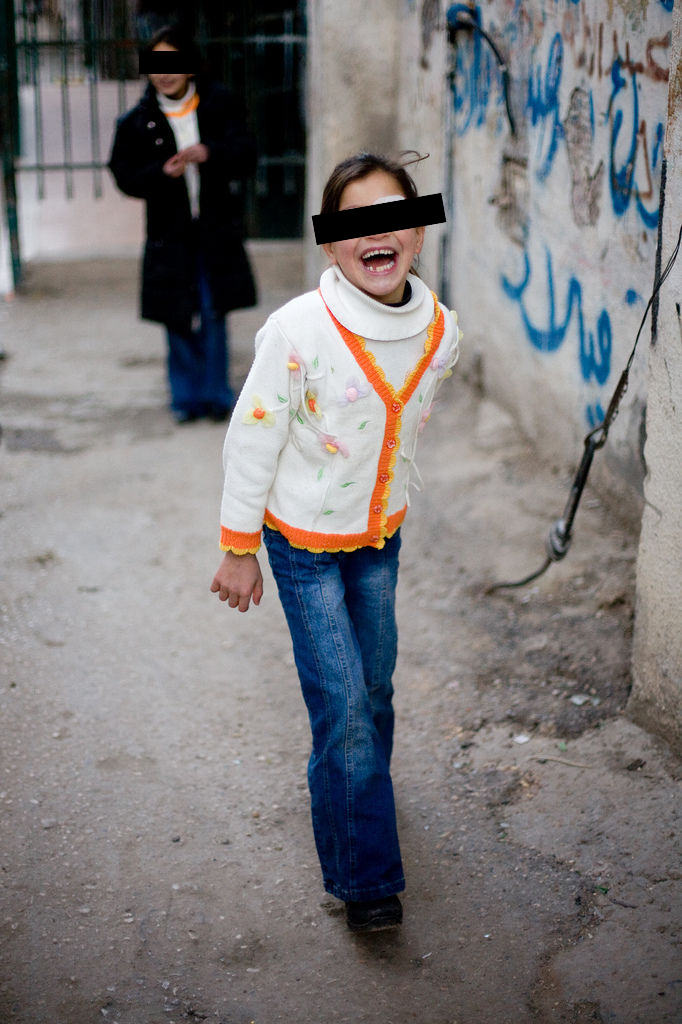 A laughing girl in a white cardigan with orange trim, eyes covered by a black bar, walks down an alley past graffiti-covered walls
