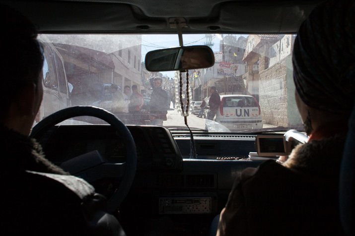 View from the back seat of a car of a street with a white UN vehicle and people crossing, prayer beads hanging from the mirror