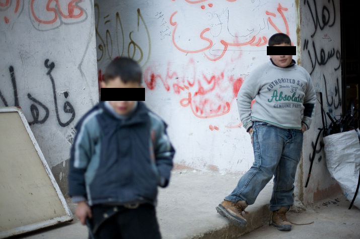 Two boys, eyes covered by black bars, in front of a wall covered in Arabic graffiti
