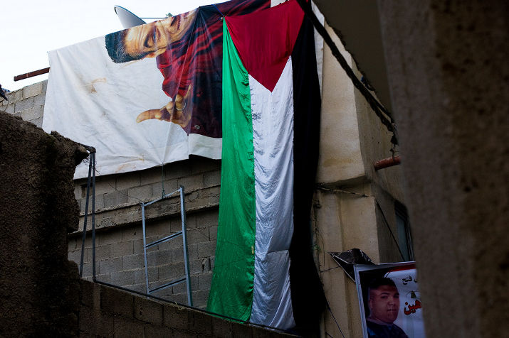 A large Palestinian flag and a banner with a man's portrait hang on a wall, with a small martyr poster below