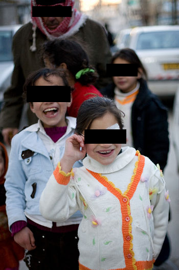 A group of children, eyes covered by black bars, laughing and posing in the street, an adult in a keffiyeh behind them