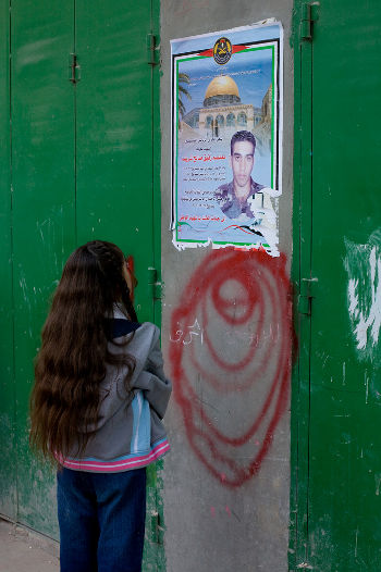 A girl with long hair looks up at a poster of a young soldier and the Dome of the Rock on a green metal door