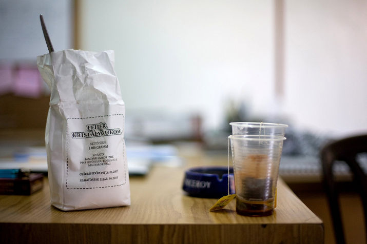 A paper bag of sugar with a spoon in it and a plastic cup of mint tea next to an ashtray on a desk