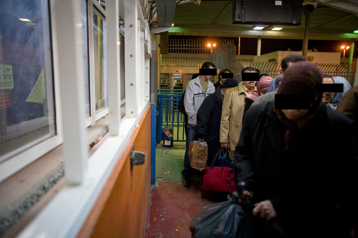 People with bags queue past a checkpoint booth at night, eyes covered by black bars