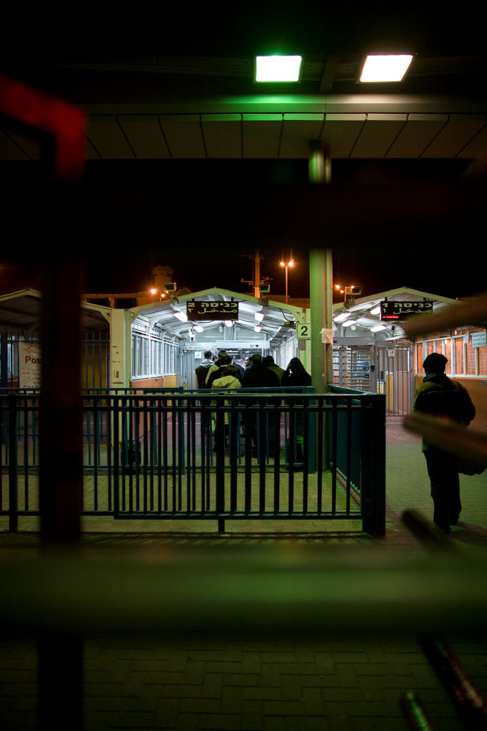 Checkpoint turnstiles at night seen through a fence, people waiting under lit shelters with Hebrew and Arabic signs