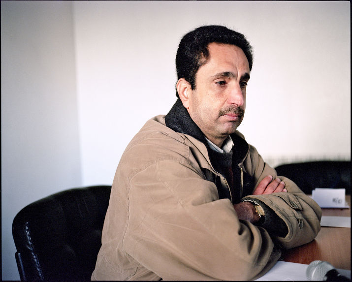 A man in a tan jacket sits with his arms crossed at a meeting table with a microphone