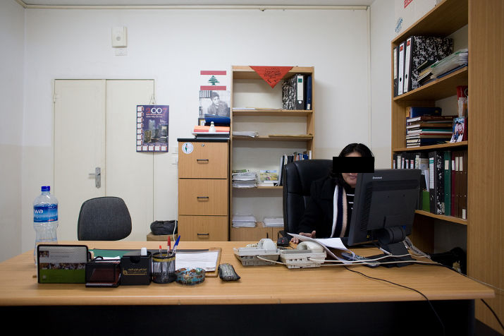 A woman smiles at a computer screen in a small office with a bookshelf of binders