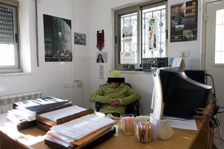 A young man in a yellow hooded sweatshirt sits behind a paper-strewn desk, with a Stop the Apartheid Wall poster on the wall