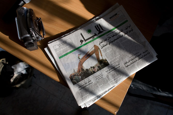 An Arabic newspaper and a camcorder on a table in slanted sunlight, the front page showing a bulldozer and a crowd
