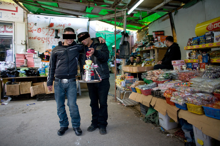Two young men, eyes covered by black bars, pose arm in arm in a market street next to a sweets stand
