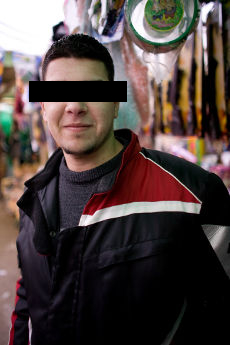 Portrait of a smiling young man in a black and red jacket, eyes covered by a black bar, in a market street