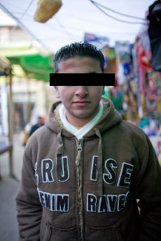 Portrait of a young man in a brown hooded sweatshirt, eyes covered by a black bar, in a market street