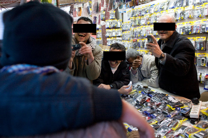 Four men laughing behind the counter of a mobile phone accessories stand, eyes covered by black bars, one filming with a phone