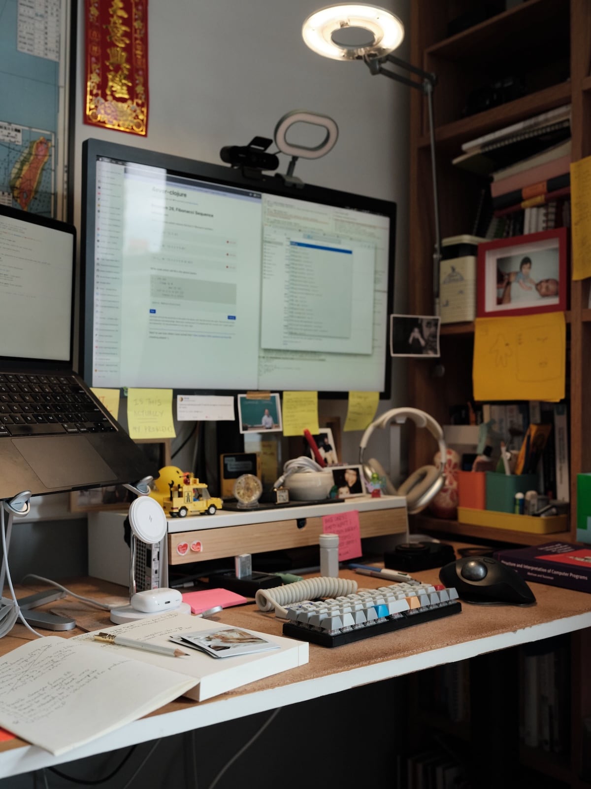 A photograph of a messy work desk, with a laptop raised ona stand, a large screen with sticky notes hanging off it, a keyboard, a trackball, headphones on a stand, books and notebooks, and various assorted items.